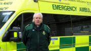 The Revd Ian Charles wearing a clerical shirt and dog collar underneath an ambulance service coat in front of an emergency ambulance